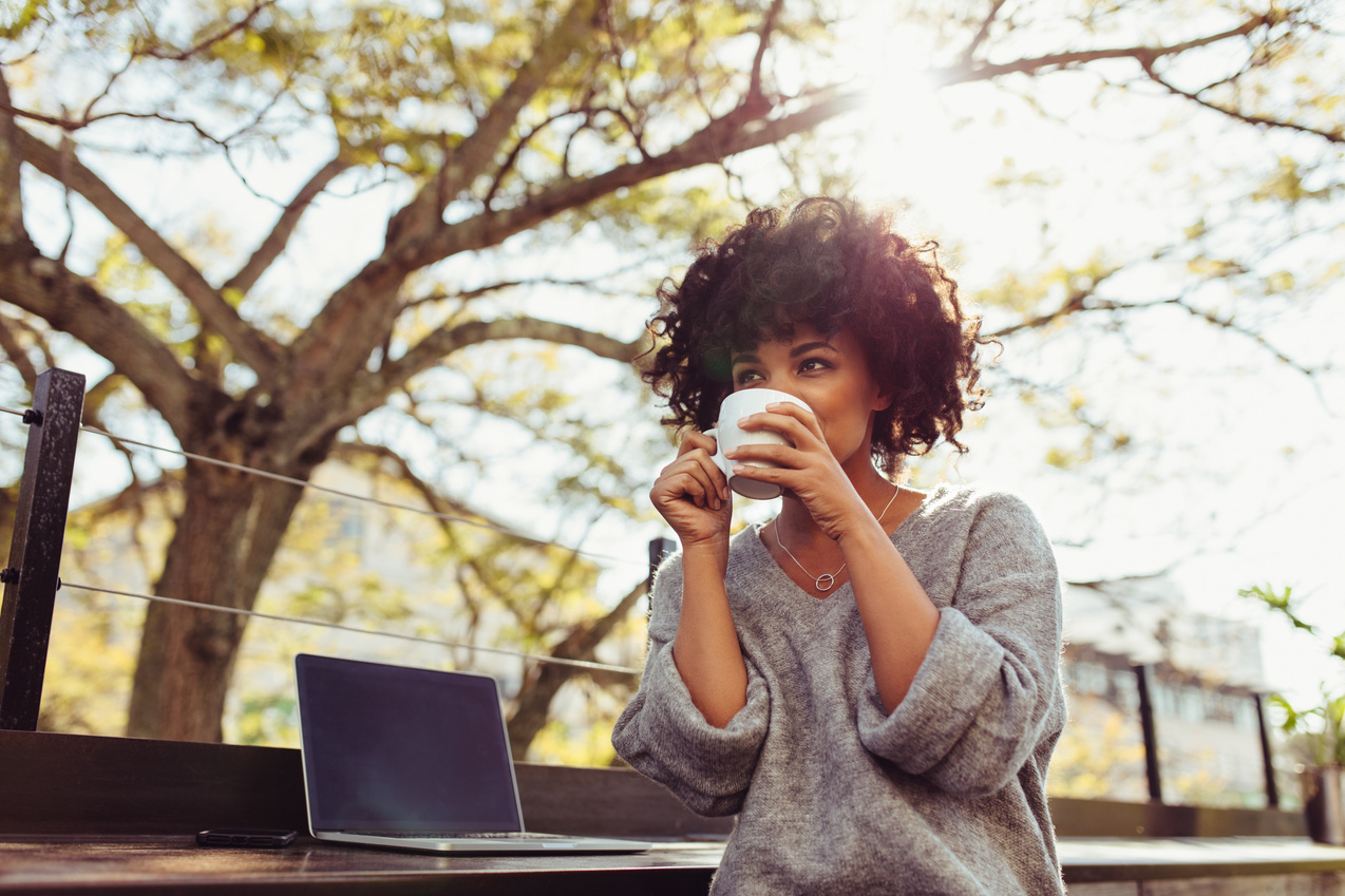 Black woman with curly afro sips coffee outside next to open laptop